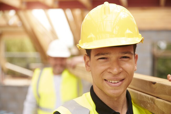 Portrait Of Builder And Apprentice Carrying Wood On Site